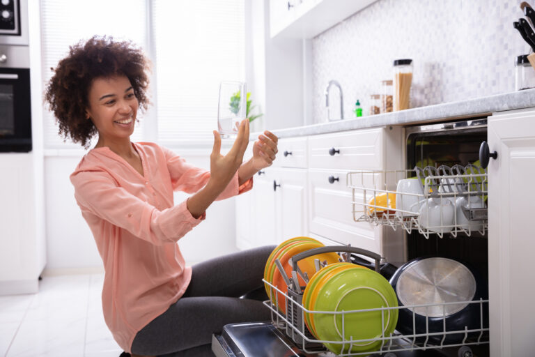 Happy Young Woman Checking Washed Transparent Drinking Glass Near The Dishwasher